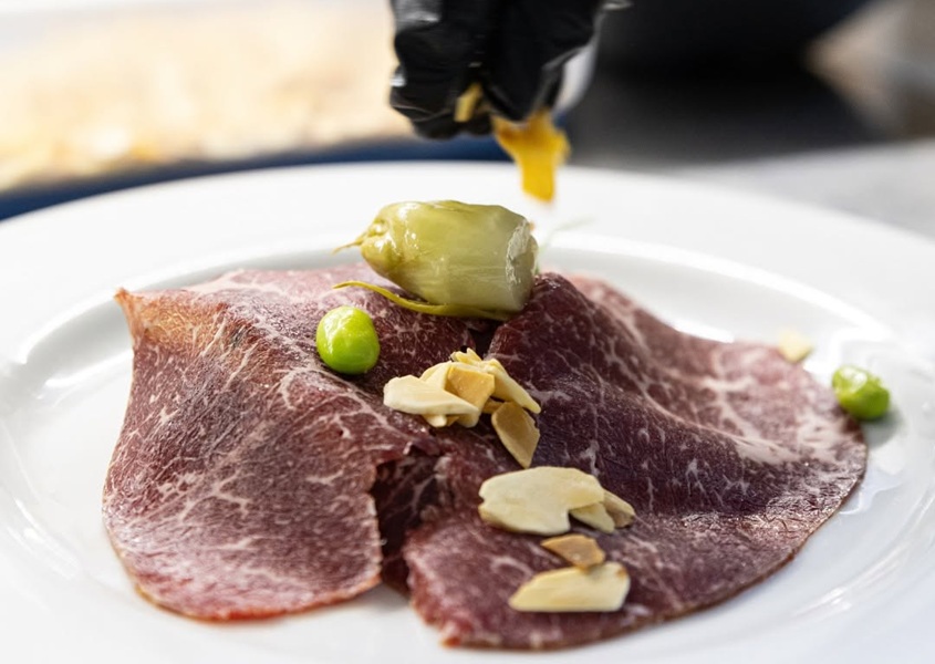 Fettine di carne marmorizzata su piatto bianco con lamelle di mandorla, piselli e peperoncino verde, mano in guanto nero